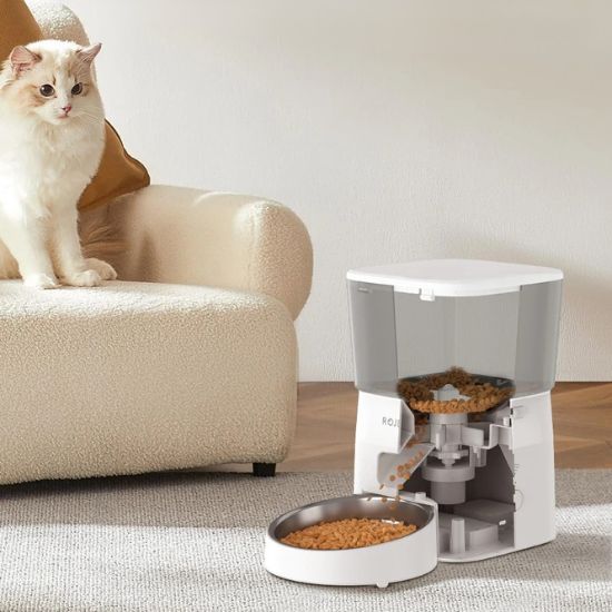 Chat blanc assis près d'un distributeur automatique croquettes chat sur un canapé, illustrant son utilisation pratique et système anti-blocage.